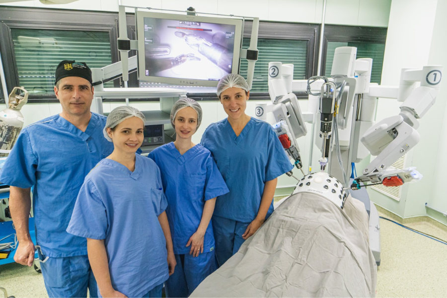 Equipe de quatro médicos sorriem ao lado de um robô cirurgião, parte do treinamento prático para que o profissional se qualifique em cirurgia robótica.
